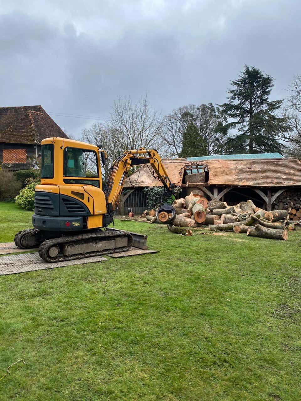 This is a photo of a tree which has grown through the roof of a barn that is being cut down and removed. There is a digger that is removing sections of the tree as well. Castle Donnington Tree Surgeons